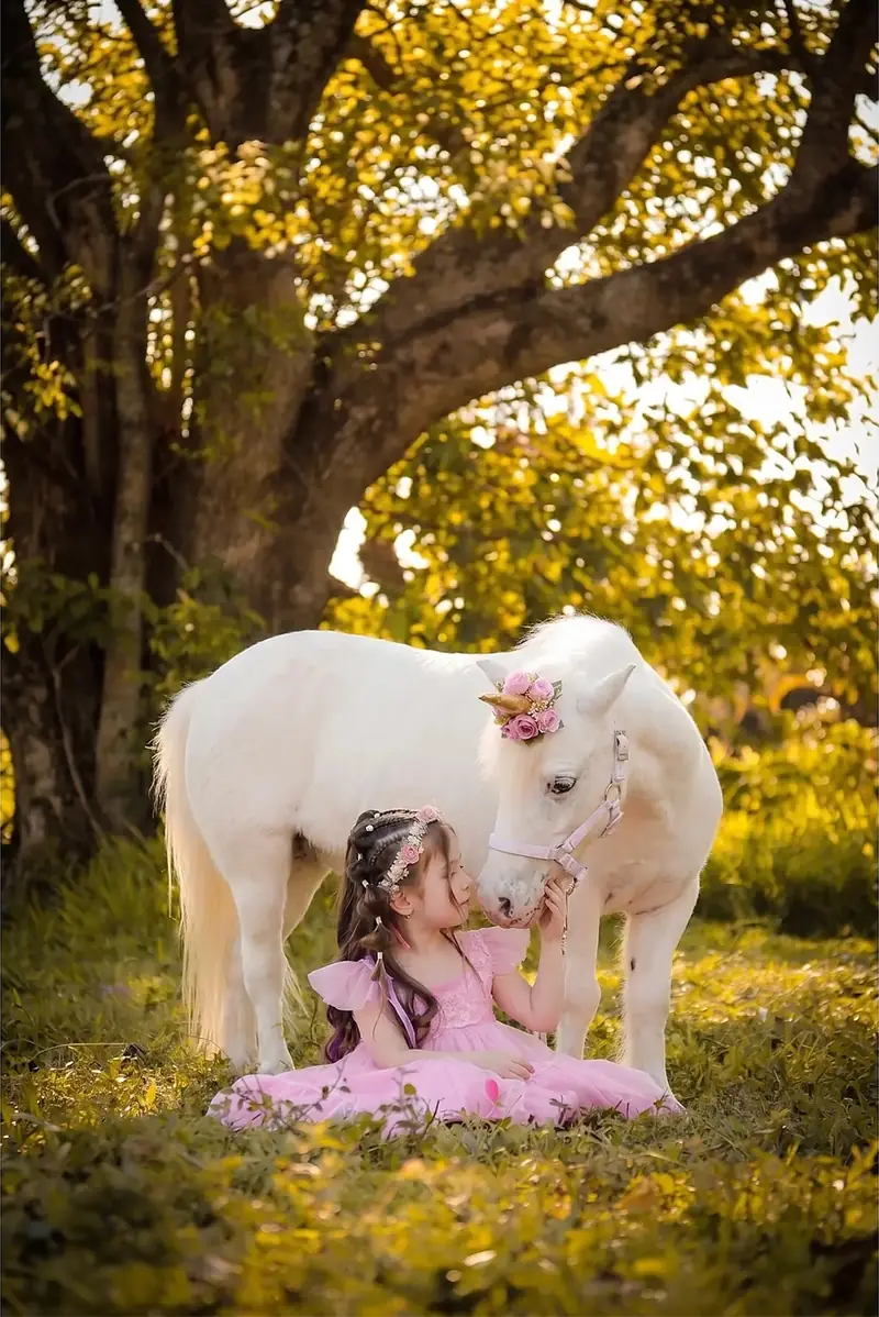 Niña sentada en el pasto junto a un poni blanco