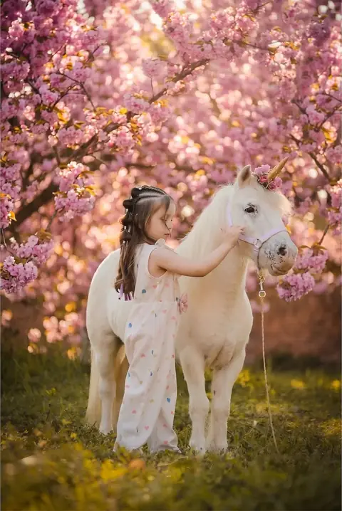 Niña acariciando a un poni blanco bajo un árbol de flores rosadas