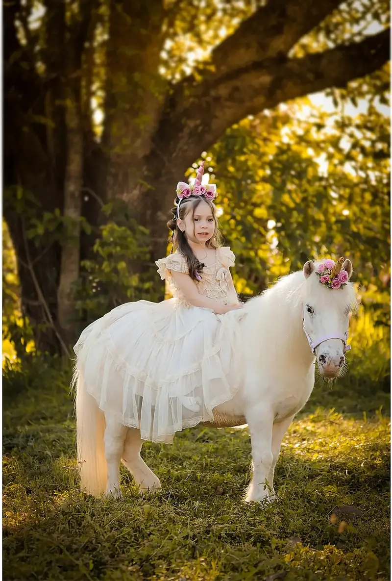 Niña de vestido blanco sentada sobre un poni blanco en el parque