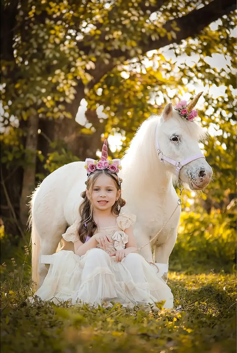 Niña con corona de flores sentada junto a un poni blanco, con luz dorada