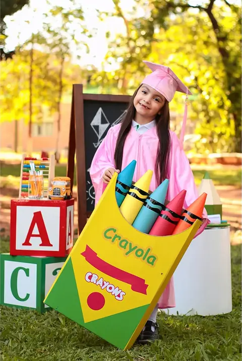Niña con toga rosada junto a una caja de crayones gigante